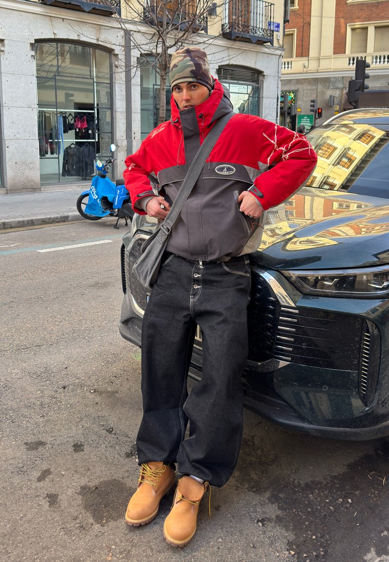 Homme portant une veste rouge et grise, un chapeau camouflage, un jean large noir et des bottes couleur cuir clair, appuyé contre une voiture noire dans une rue urbaine.