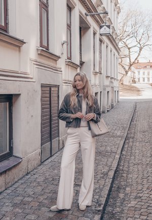 Mujer con cabello largo y rubio que lleva una chaqueta gris y pantalones blancos, de pie en una calle empedrada junto a edificios blancos.