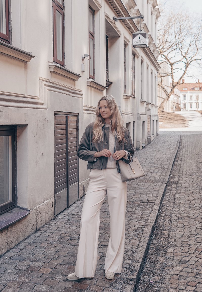 Femme aux longs cheveux blonds portant une veste grise et un pantalon blanc, debout sur une rue pavée à côté de bâtiments blancs.