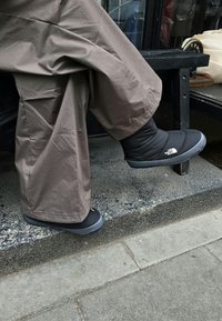 Sorte isolerede støvler med en tekstureret stofoverdel og grå gummisåler. Båret med løse beige bukser, sat mod en betonbaggrund.