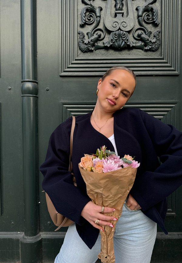 Un bouquet de fleurs roses et crème enveloppé dans du papier brun. Le modèle porte une veste oversize noire et un jean bleu clair, se tenant devant une porte vert foncé.