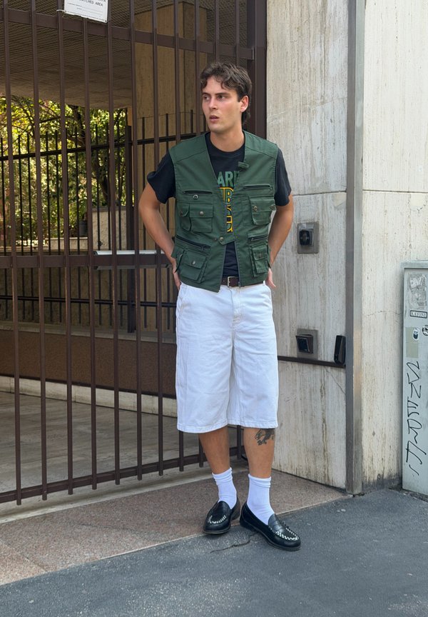 Jonge man met kort donker haar die een groene gilet draagt, een zwart T-shirt, witte knielange short, witte sokken en zwarte schoenen en die naast een poort staat.