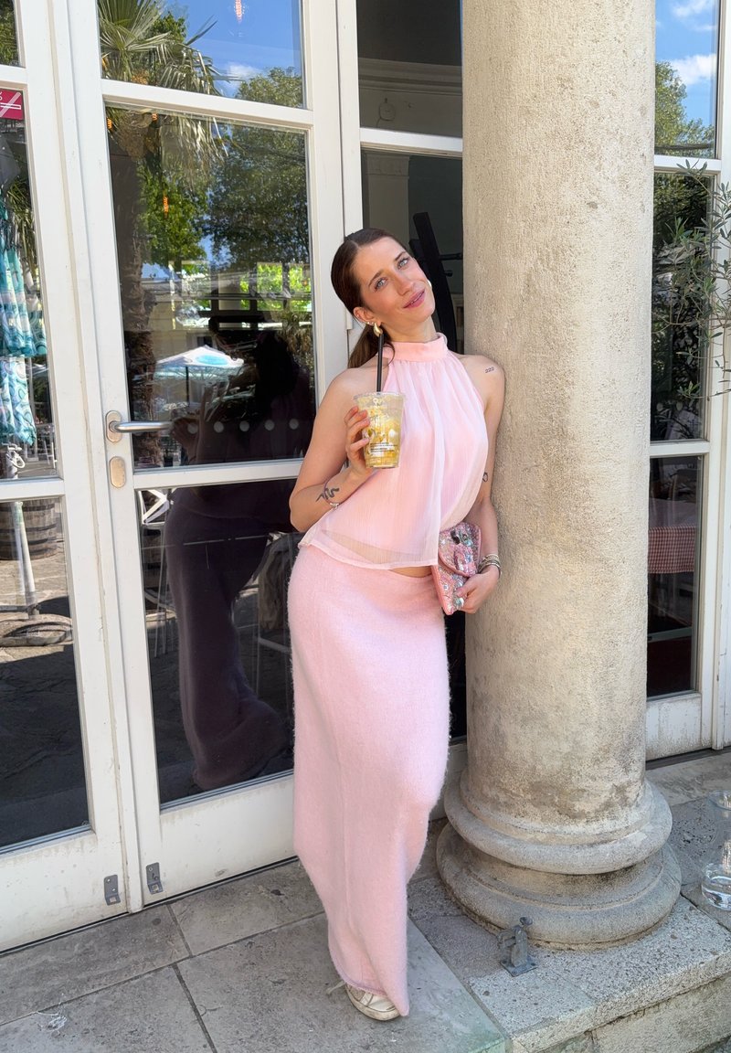 Femeie îmbrăcată într-un top pal roz deschis fără mâneci și o fustă lungă, ținând o băutură și o poșetă, sprijinită de o coloană mare de piatră lângă uși de sticlă.