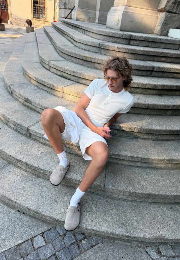 Jeune homme en chemise blanche, short et pantoufles, assis sur des marches en pierre courbées en extérieur près d’un bâtiment, regardant vers le bas sur le côté.