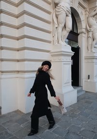 Černý trenčkot s páskem, černé kozačky nad kolena a dvoubarevná kabelka. Stojí na kamenné dlážděné ulici poblíž zdobné bílé architektury.