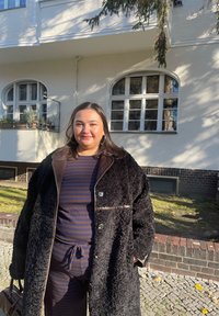 Zwarte gelaagde jas met een bruine kraag en knoopsluiting; gedragen over een marineblauw en bruin gestreepte outfit, voor een lichtgekleurde gevel.