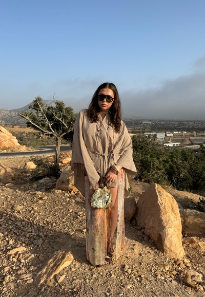 Kaftán beige claro con elegante bordado, combinado con un bolso estampado. Los tonos tierra y un diseño fluido complementan el entorno natural.
