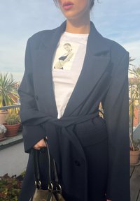 Marineblauwe lange jas met een dubbelzijdig ontwerp en riem; gelaagd over een wit t-shirt met een monochrome grafiek. Geweven stof met zakken.