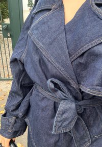 Jeansjacke mit Wickeldesign, mit einem taillierten Bund, breiten Revers und kontrastierenden Nähten. Die Farbe ist ein tiefes Indigo-Blau.