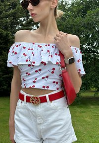 Top bianco off-shoulder con stampa di ciliegie rosse, abbinato a pantaloni corti bianchi a vita alta e una cintura in pelle rossa con fibbia oro. Borsa rossa.