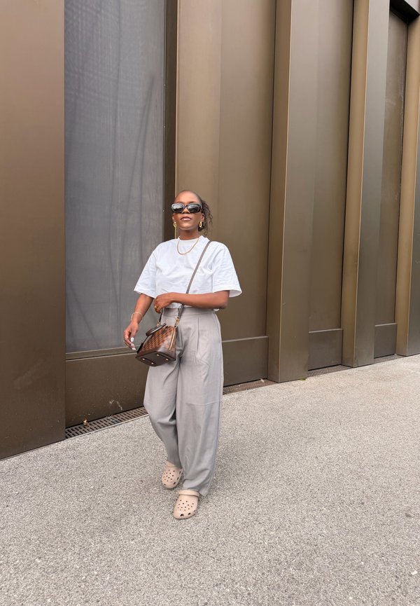Frau trägt weisses Hemd, graue weite Hose, Sonnenbrille und beige Clogs, geht auf grauem Gehweg an einer braunen modernen Gebäudewand entlang.