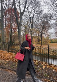 Ženska oblečena v črno dvoredni plašč in rdeč šal, drži roza torbico, hodi po poti prekriti z listjem ob ribniku.