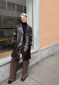 Personne souriante aux cheveux blonds portant un manteau en cuir brun foncé, un col roulé noir, un pantalon large marron et des chaussures noires à bout carré, debout à l'extérieur.