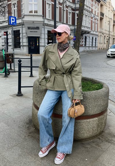 Trench coat de un verde oliva claro con cinturón, superpuesto sobre un suéter rosa, combinado con jeans acampanados de un azul claro y zapatillas burdeos. Pequeño bolso marrón.