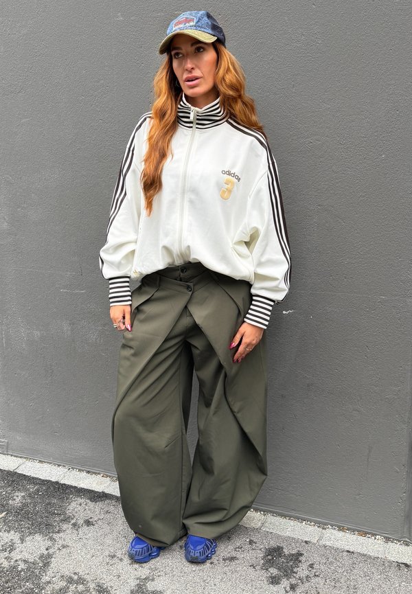 Stojąca kobieta z długimi rudymi włosami, ubrana w białą kurtkę Adidas, szerokie oliwkowe spodnie, niebieskie buty i jeansową czapkę na tle szarej ściany.