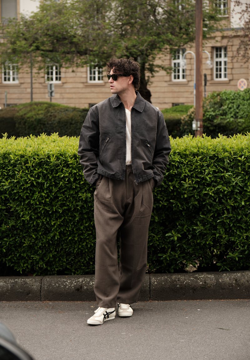 Άνδρας με σγουρά μαλλιά, που φοράει γυαλιά ηλίου, σκούρο μπουφάν, καφέ φαρδιά παντελόνια και λευκά αθλητικά παπούτσια, στέκεται στο πεζοδρόμιο κοντά σε ένα πράσινο φράχτη.