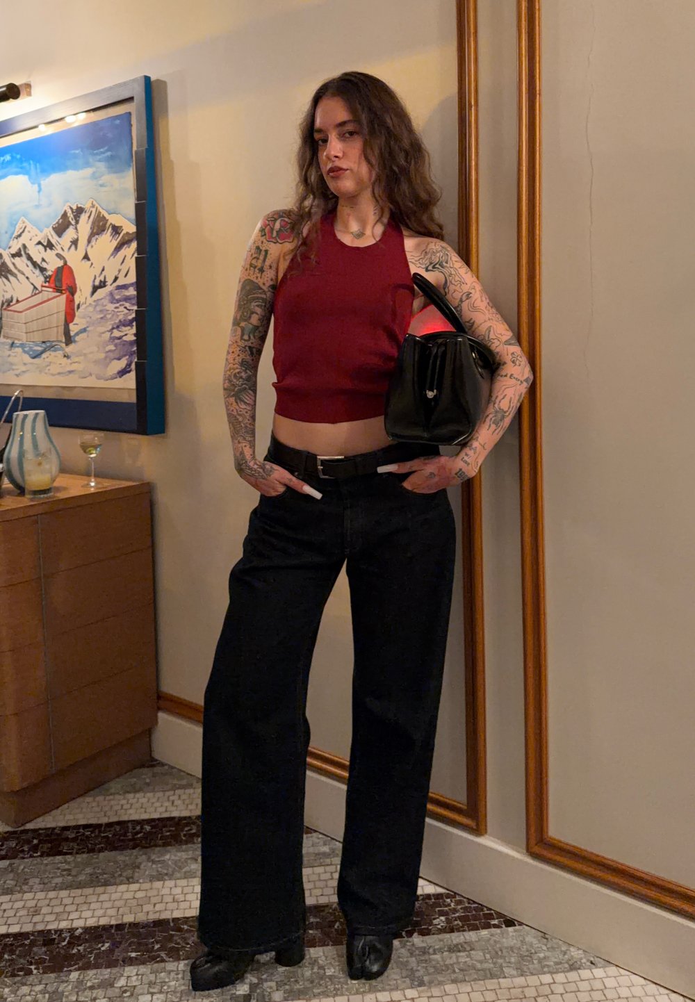 Jeune femme aux longs cheveux bouclés portant un haut sans manches rouge et un pantalon noir, tenant un sac à main noir, debout dans une pièce avec des œuvres d'art.