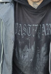 Fekete grafikás póló, amely részletes, sötét nyomatot ábrázol lándzsás, kapucnis figurákkal, fekete bőrdzsekivel párosítva.