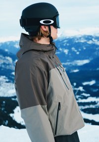 Personne de profil portant un casque de ski noir, des lunettes de protection et une veste d'hiver bicolore gris, se tenant devant un paysage de montagne enneigée.