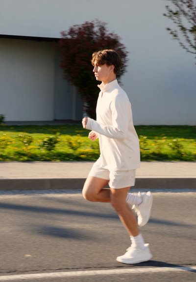 Joven corriendo en una calle pavimentada con una camiseta blanca de manga larga, pantalones cortos blancos y zapatillas blancas a la luz del día.