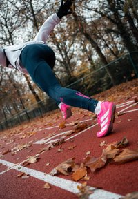 Chaussures de sport rose vif avec des bandes blanches, portées sur une piste de course ornée de feuilles d'automne, associées à des leggings bleu marine.