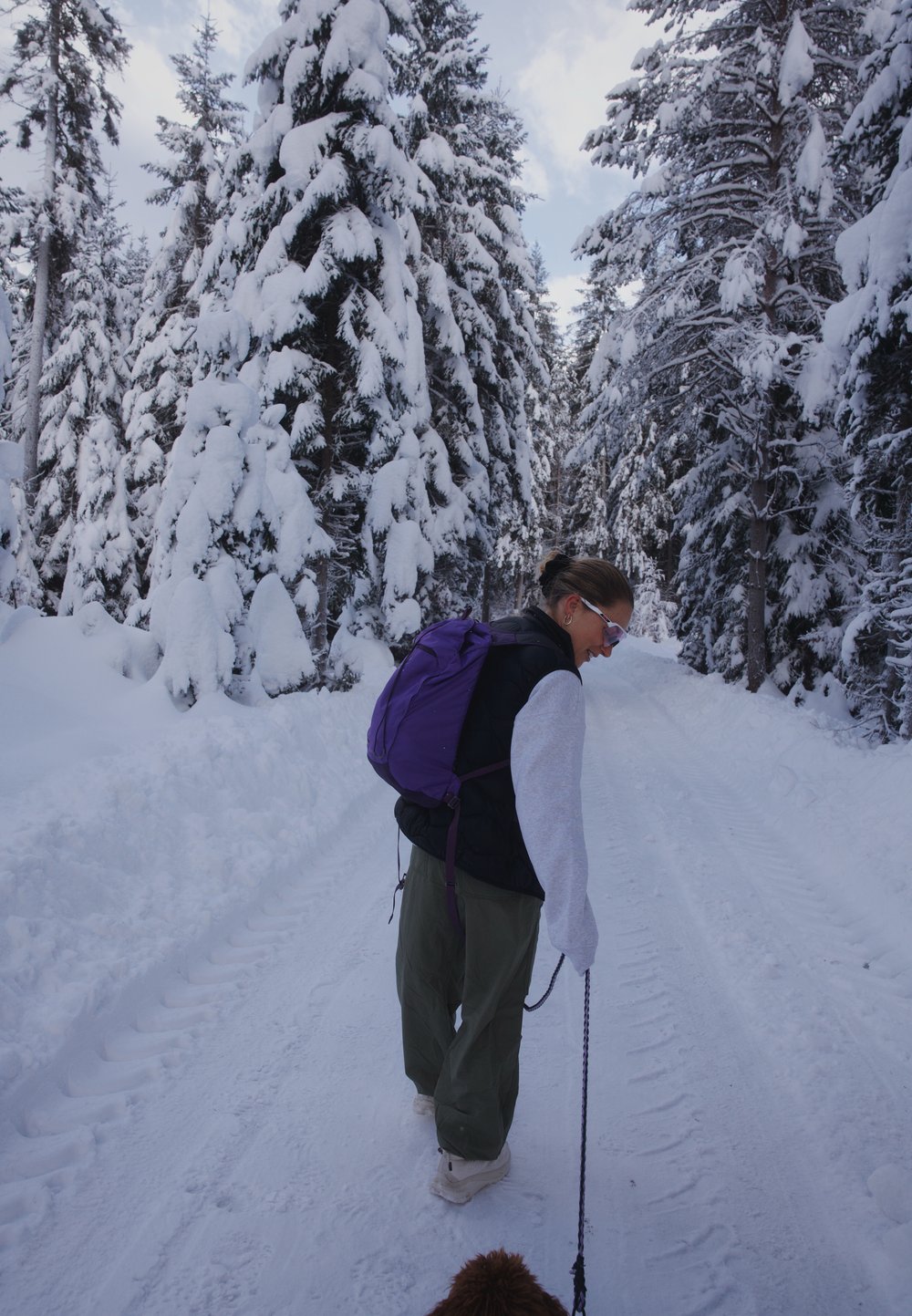 Person walking along a snow-covered path, dressed in a black padded vest, a grey sweatshirt, olive green trousers, and white boots, while carrying a purple backpack.