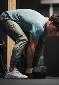 Vyras su tatuiruotėmis ir turkio spalvos marškiniais laiko hantelius, ruošdamasis kilti svorius sporto salėje.