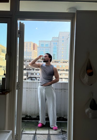 Hombre con camiseta sin mangas gris y pantalones deportivos bebiendo de una taza blanca en un balcón con edificios urbanos al fondo.