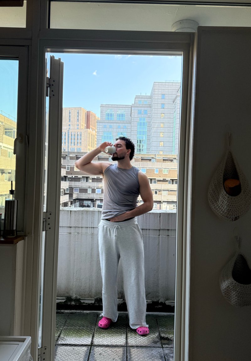 Homme portant un débardeur gris et un pantalon de survêtement, buvant dans une tasse blanche sur un balcon avec des bâtiments urbains en arrière-plan.