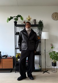 Veste en cuir noire avec un col en peau lainée et des poches, portée sur un col roulé beige clair, associée à un pantalon noir ample et des chaussures.