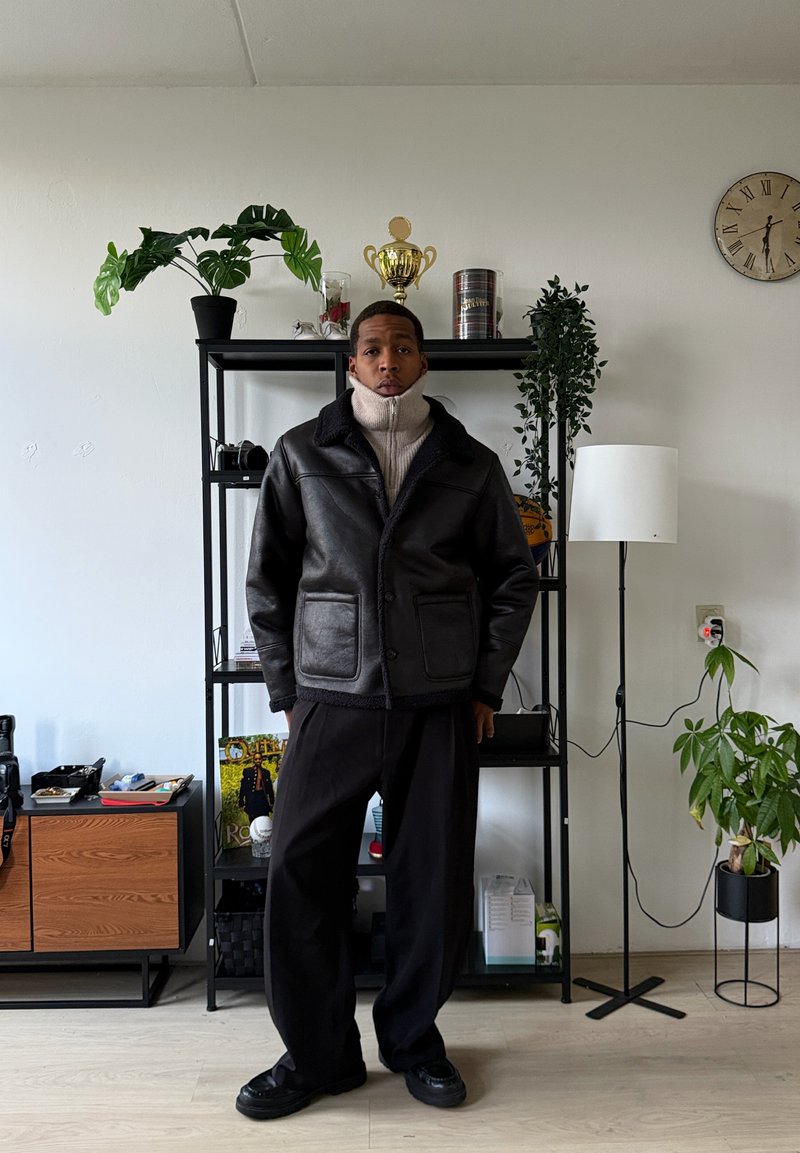 Veste en cuir noire avec un col en peau lainée et des poches, portée sur un col roulé beige clair, associée à un pantalon noir ample et des chaussures.