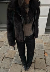 Svart konstläderjacka över en grå polokrage-tröja, i kombination med mörk denimjeans och svarta spetsiga klackar på en stenbelagd trottoar.