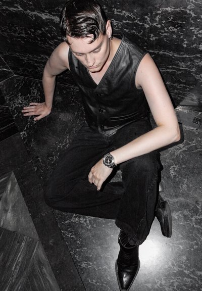 Chaleco de cuero negro sobre una camiseta sin mangas, combinado con jeans negros y botas negras de punta. Se lleva un reloj plateado en la muñeca izquierda.