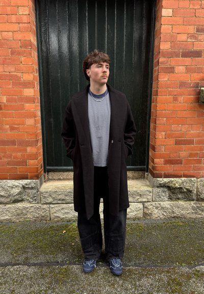 Abrigo negro sobre una camisa a rayas, pantalones anchos oscuros y zapatos azul con estampado; de pie contra una pared de ladrillo texturizada y una puerta de madera.