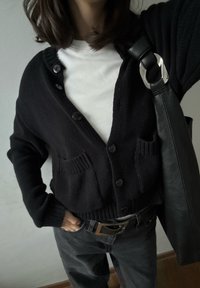 Personne portant un cardigan noir boutonné sur une chemise blanche, un jean sombre avec une ceinture, et un sac en cuir noir porté à l'épaule.