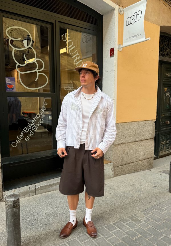 Joven con gorra marrón, camisa blanca de rayas, pantalones cortos oscuros y zapatos marrones, está en la acera de la ciudad frente a una cafetería.