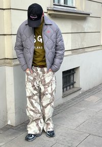 Casaco cinza acolchoado, sweatshirt verde-oliva com letras brancas, calças cargo estampadas em creme claro e castanho, e sapatilhas pretas.