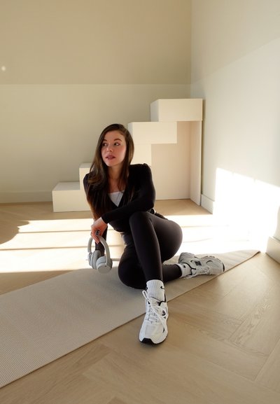 Camiseta negra de manga larga, leggings negros y zapatillas gris. Auriculares gris claro sostenidos en una mano. Suelo de madera natural y paredes color crema.