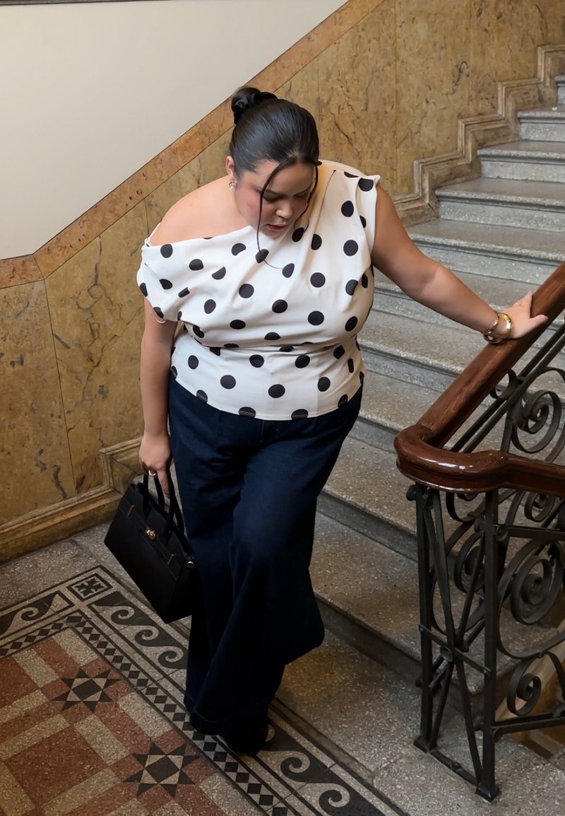 Moteris vilki baltą marškinėlį su dėmėmis ir tamsias kelnes, laiko juodą rankinę ir lipa akmeninėmis laiptais su mediniu turėklu.