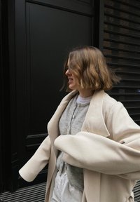 Femme aux cheveux courts et ondulés portant un manteau beige et un pull gris, debout de profil sur un fond noir, souriante et bougeant les bras.