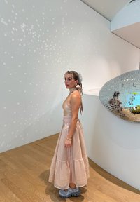 Saia maxi em camadas, de cor rosa claro, combinada com uma blusa justa bege. Apresenta um tecido texturizado, com um laço na parte de trás e calçado cinza.