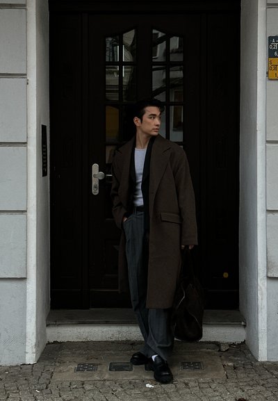 Un hombre está junto a una puerta de madera oscura, vestido con un abrigo marrón, pantalones grises y una camisa gris clara, sosteniendo una bolsa oscura.