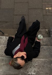 Personne assise sur des marches en béton portant un manteau noir et des lunettes de soleil, tenant un smoothie vert avec une paille, écharpe écarlate visible.