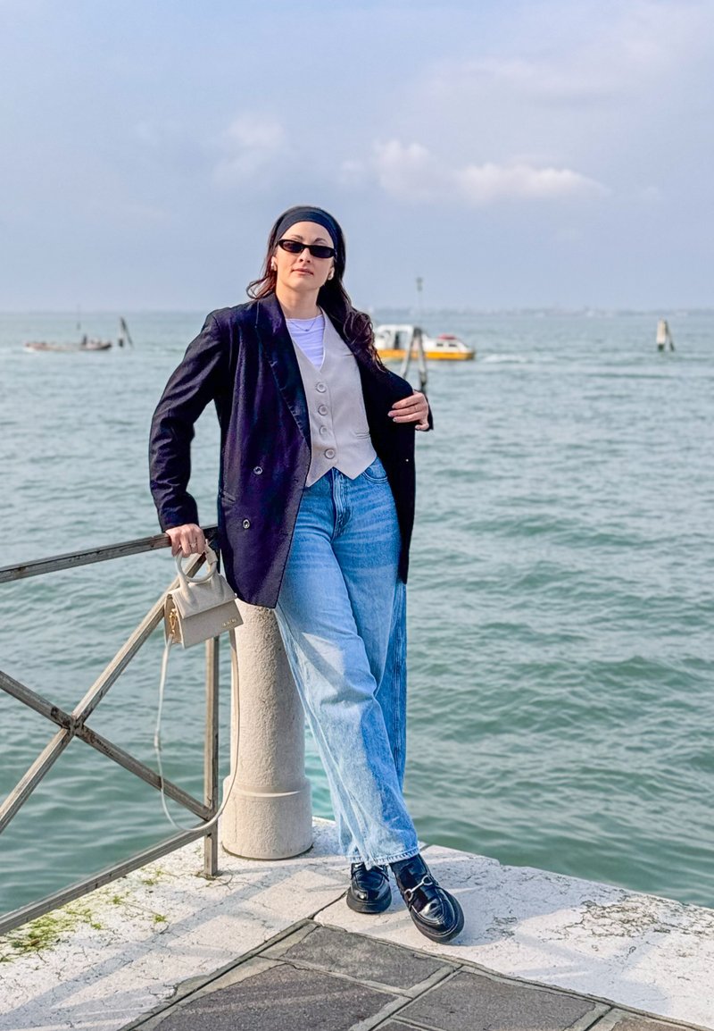 Femme portant des lunettes de soleil, un blazer noir, un gilet beige et un jean, appuyée sur une rambarde près de l'eau avec des bateaux et un ciel nuageux en arrière-plan.