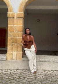 Haut à halter marron avec tissu drapé, pantalons larges crème et accessoires dorés. Colonne en pierre en arrière-plan avec un pavé texturé.