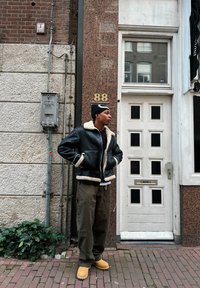 Fekete shearling dzseki krém béléssel, sötét cargo nadrág és barna csizma, egy téglafal és egy fekete akcentusokkal díszített fehér ajtó előtt áll.