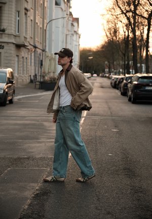Joven con ropa casual y gorra caminando por una calle tranquila de la ciudad bordeada de coches aparcados al atardecer.