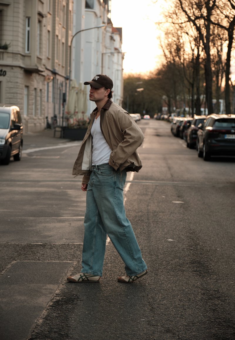 Jeune homme en vêtements décontractés et casquette traversant une rue calme de la ville bordée de voitures stationnées au coucher du soleil.