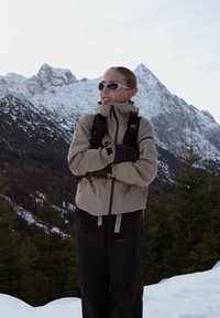 Personne portant des lunettes de soleil et des vêtements d'hiver, debout les bras croisés dans un paysage montagneux enneigé avec des pins.