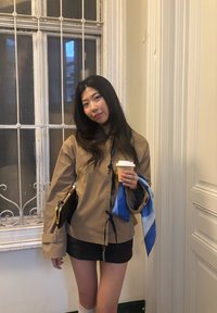 Jeune femme en veste beige et jupe noire tenant une tasse de café et une écharpe bleue, debout à l'intérieur près d'une fenêtre avec des barreaux métalliques.